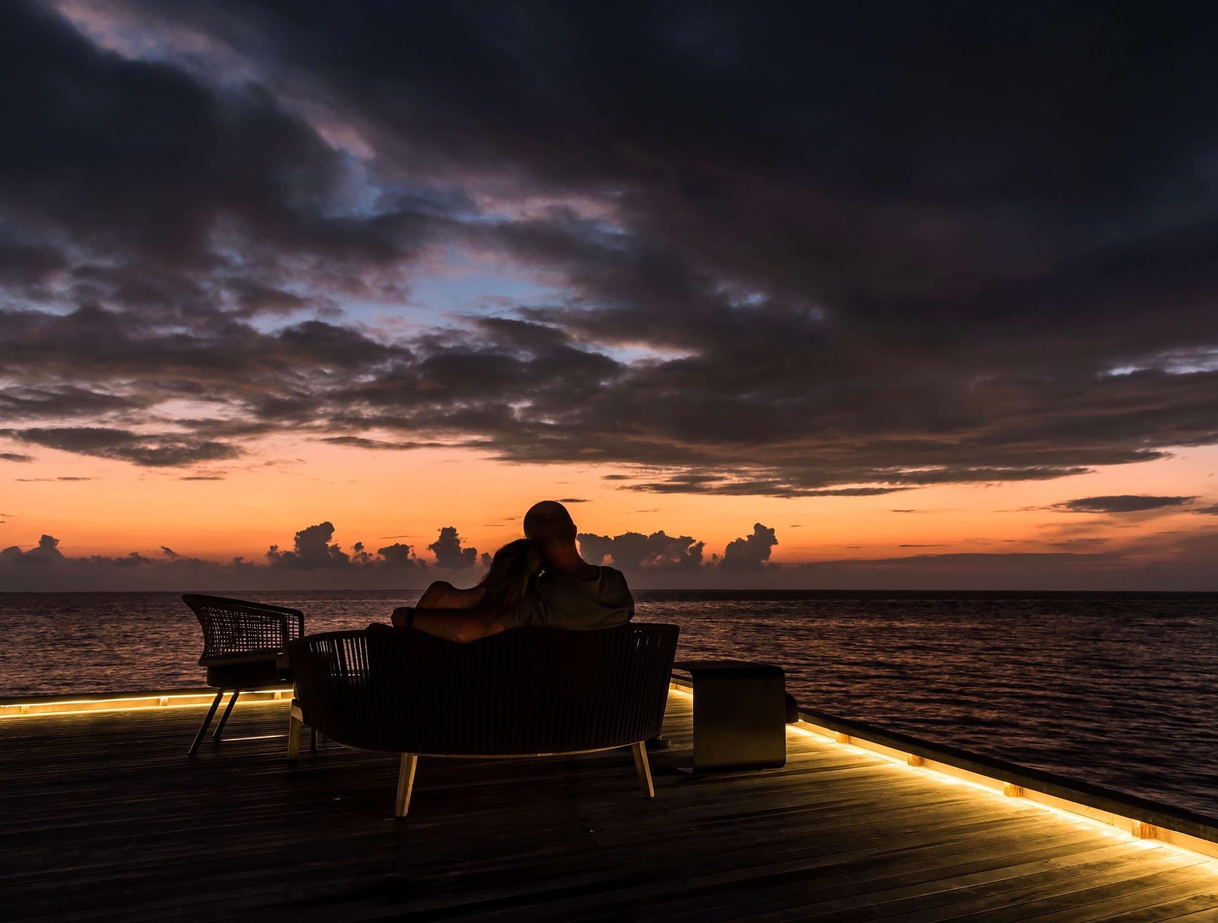 Two people sitting close together on an overwater deck, watching the sunset.